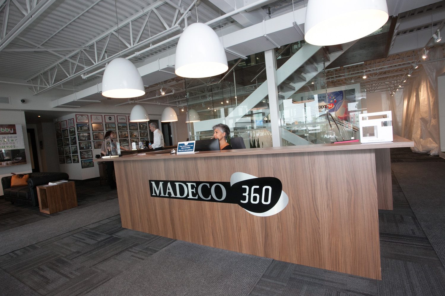 L’accueil des bureaux Madeco 360 : comptoir de bois sous des suspensions blanches, mur de cadres des projets derrière