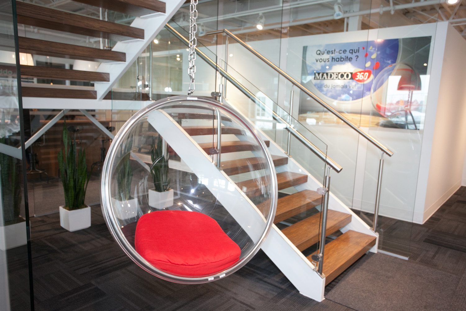 Le fauteuil bulle suspendu des bureaux Madeco : coussin rouge, escalier de bois et de verre, murale Madeco 360 derrière