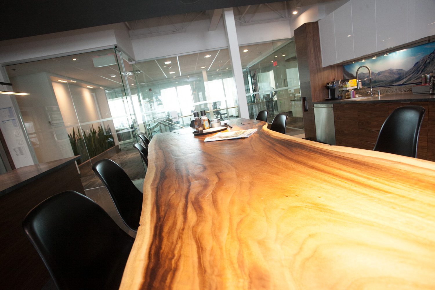 La table de conférence taillée dans une tranche de bois massif, chaises noires, coin café et salles vitrées derrière